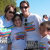 Four people wearing matching white The Color Run shirts and race bibs pose together outdoors, smiling at the camera. Two adults stand behind two children. Trees and other participants are visible in the background.