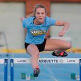 A female athlete in a blue Marquette shirt and black shorts leaps over a hurdle on an outdoor track during a race.