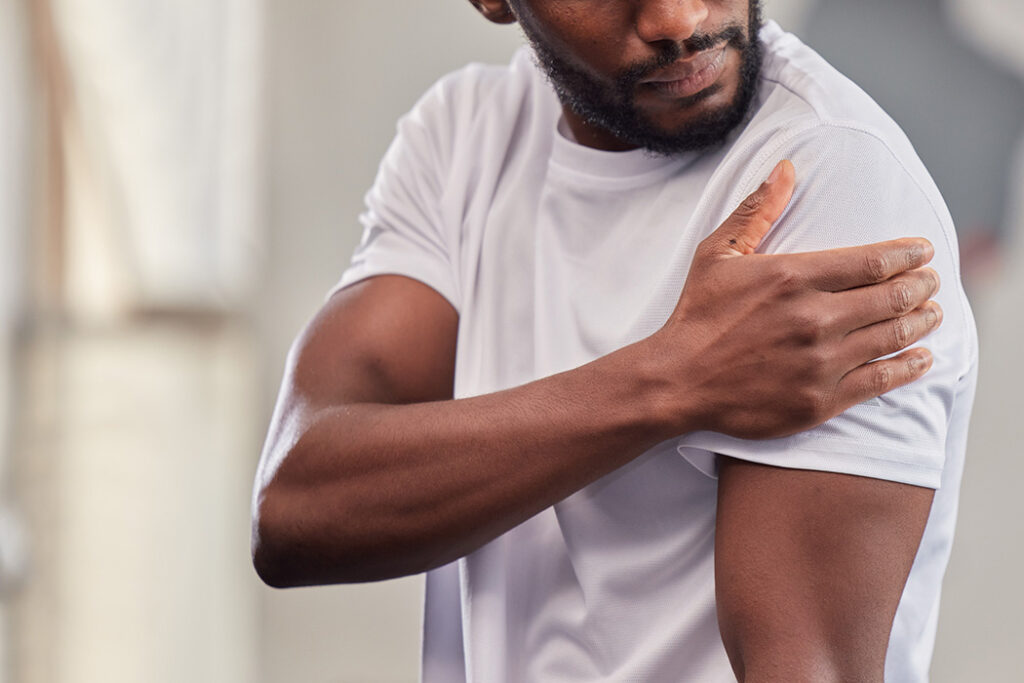 A man wearing a white t-shirt holds his left shoulder with his right hand, appearing to be in discomfort or pain. The background is blurred.