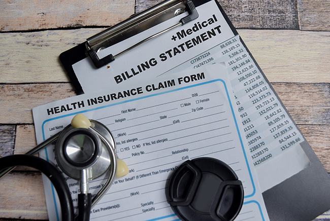 A stethoscope and lens cap rest on a health insurance claim form, with a medical billing statement on a clipboard in the background, all on a wooden surface.