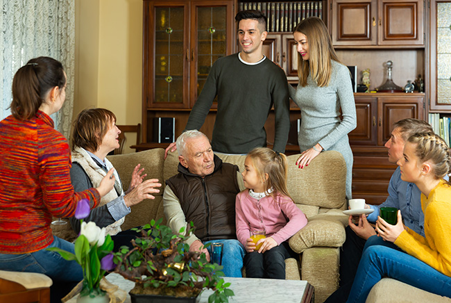 A multigenerational family gathers in a cozy living room, sitting and standing around a couch, smiling and talking together. Two children sit with adults, and some people hold cups, creating a warm, friendly atmosphere.
