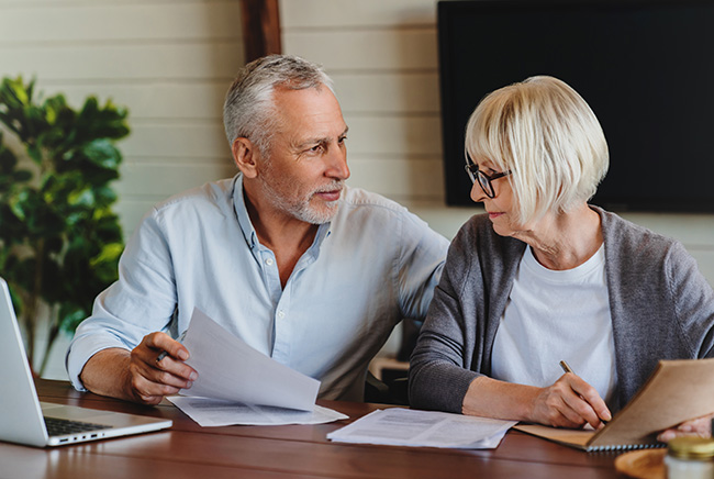 An older man and woman sit at a table with papers, a notebook, and a laptop. The man holds documents and looks at the woman, who is writing in the notebook. They appear to be discussing something together.