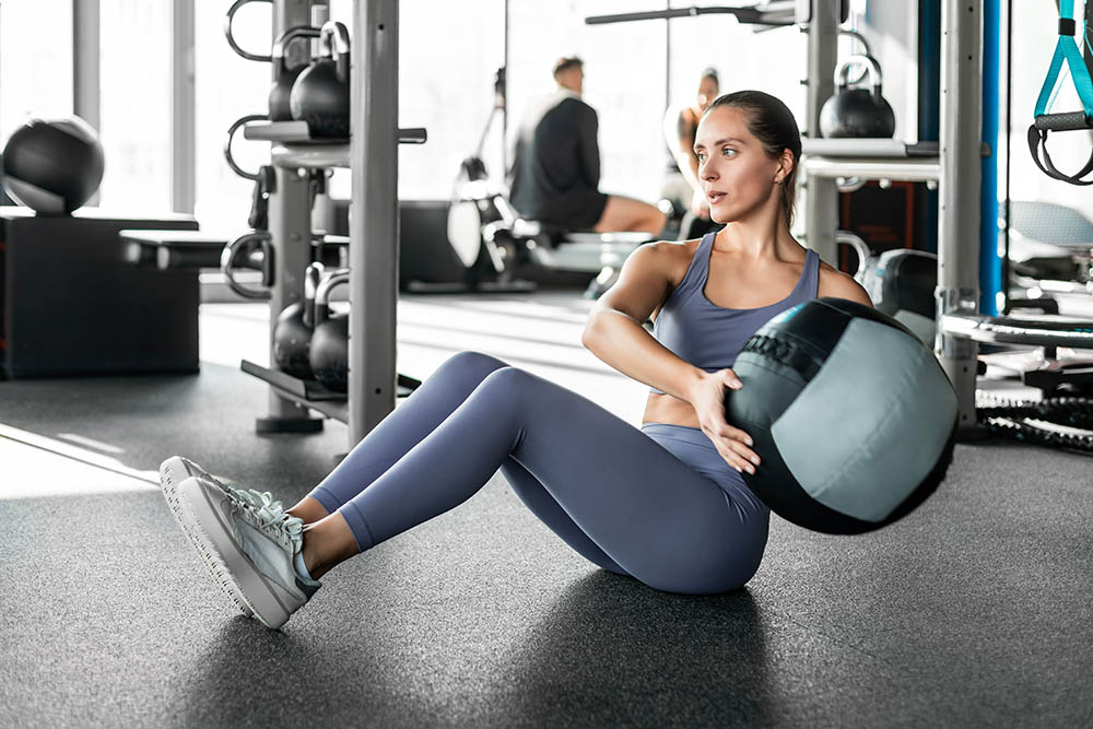 A woman in athletic wear sits on a gym floor, holding a medicine ball and twisting her torso to exercise. Gym equipment and other people are visible in the background.