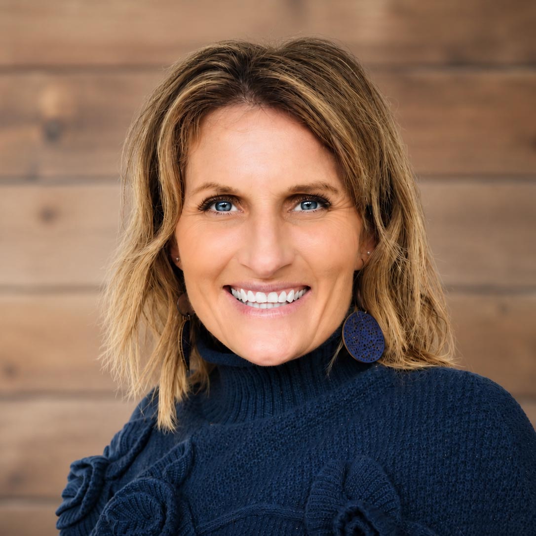 A woman with light brown hair and blue eyes smiles, wearing a navy blue sweater and dark earrings. The background is blurred wood paneling.
