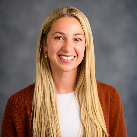 A young woman with long blonde hair smiles at the camera. She is wearing a white top and a brown cardigan, with a neutral gray background behind her.